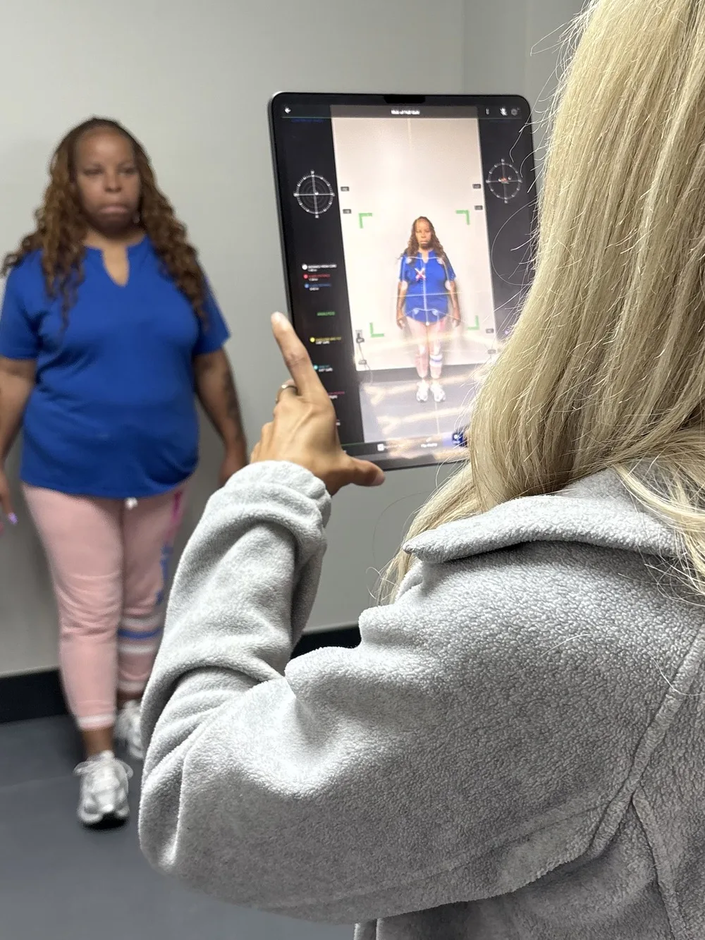 Clinician holding a tablet running Kinetisense [Gait Analysis for Fall Analysis] markerless motion capture with joint tracking markers and center-of-mass crosshairs overlaid on a patient standing for postural and gait assessment at Padda Institute, St. Louis