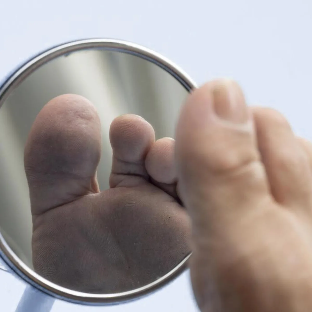 Hand holding a round mirror to inspect the sole of a foot, the daily self-check recommended when protective sensation is reduced by peripheral neuropathy