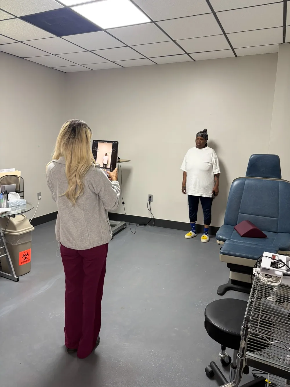 Clinician holding a tablet running Kinetisense [Gait Analysis for Fall Analysis] markerless motion capture with joint tracking markers and center-of-mass crosshairs overlaid on a patient standing for postural and gait assessment at Padda Institute, St. Louis